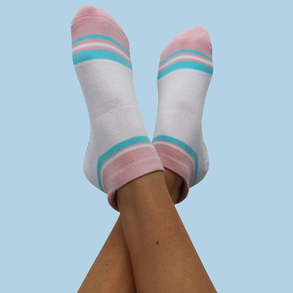 Grip-Socks-Calypso-short A woman's legs in cozy white and pink socks featuring blue stripes, casually positioned on a light-colored background.